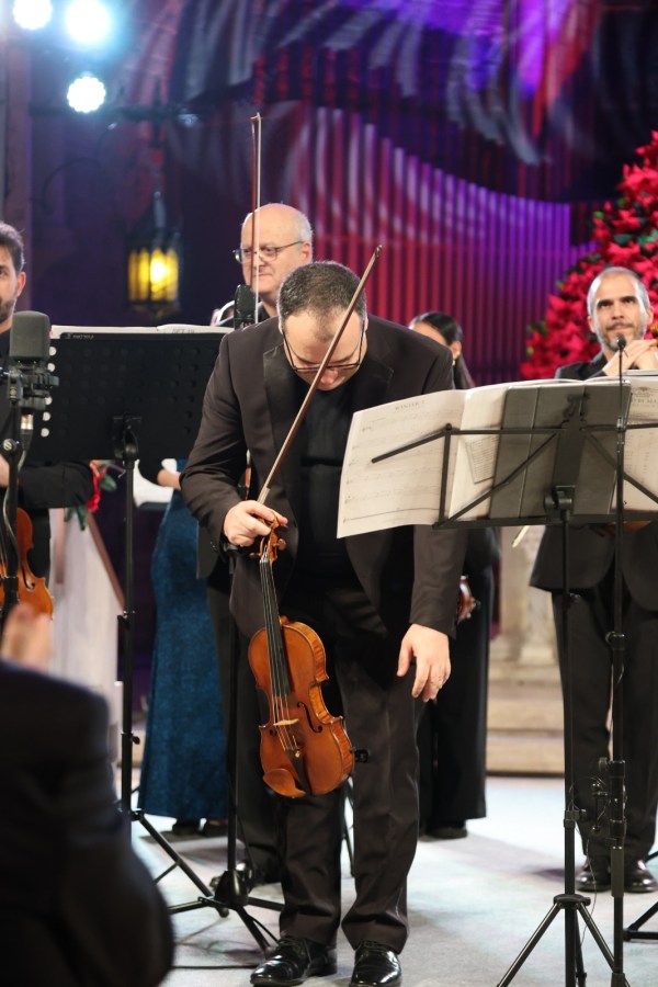 Musician bowing with a violin on stage, surrounded by an orchestra and colorful backdrop, during a performance.