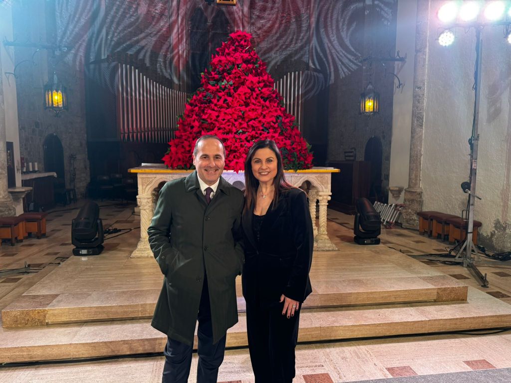 Two individuals pose for a photo in a church setting, with a large display of red flowers in the background.