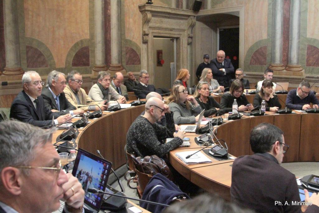 A group of people seated at a large circular table in a conference room, attentively engaging in discussion. Some individuals are using laptops and smartphones while others listen intently.