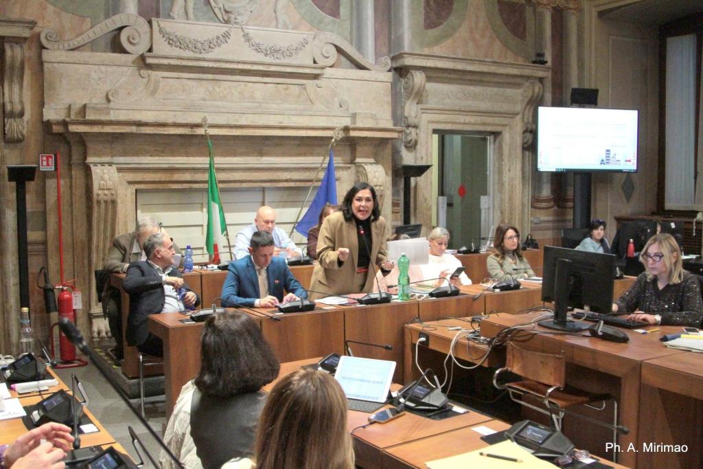 A group of people engaged in a meeting, with one woman speaking animatedly at the front and several others seated at tables with laptops and documents. The room features classical architecture and flags.