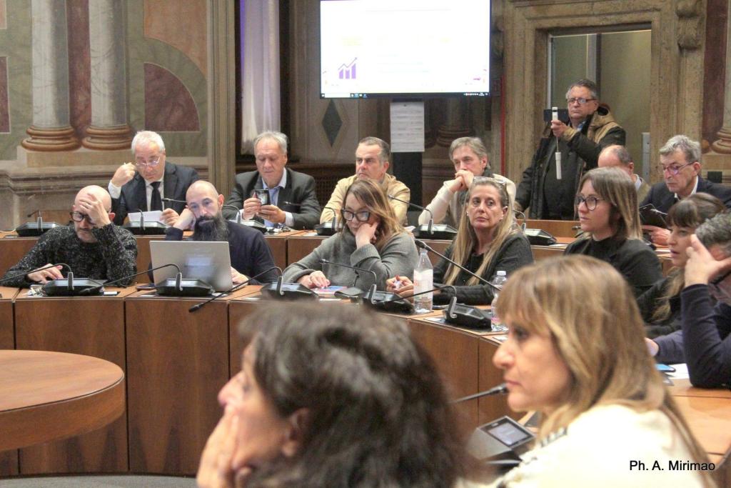 A group of people seated at a conference table during a meeting, with some individuals looking at screens and others engaged in discussion.