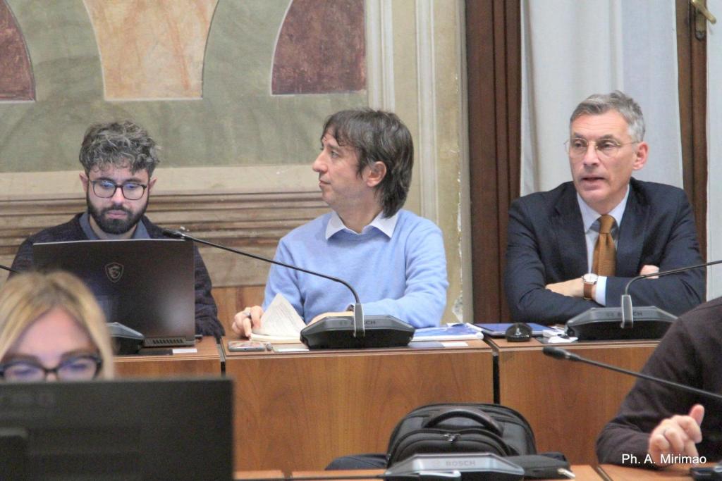 Three men seated at a table in a meeting room, with a laptop and paperwork in front of them. One man has curly hair and glasses, while another wears a light blue sweater. The third man is dressed in a suit.