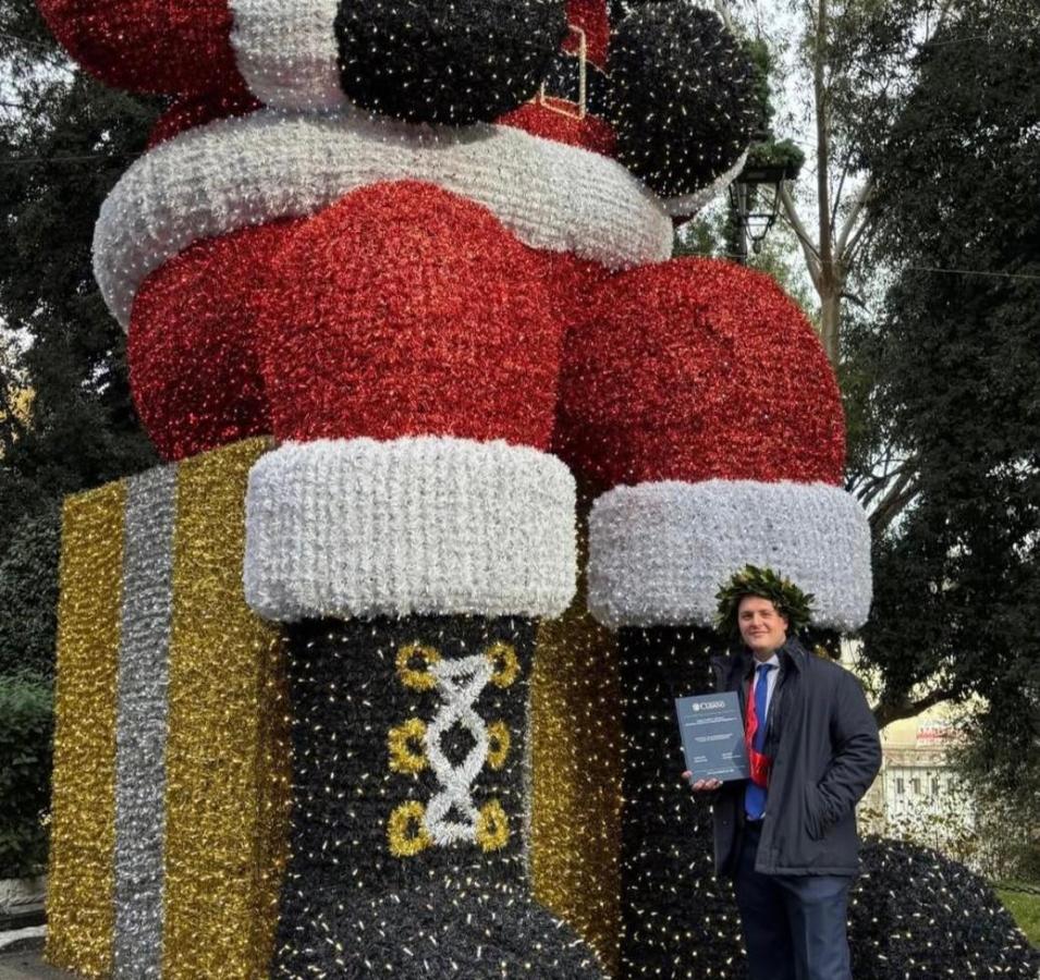 Un uomo con una corona di alloro posa accanto a una grande scultura di Babbo Natale decorata con luci colorate, mentre tiene un certificato in mano.