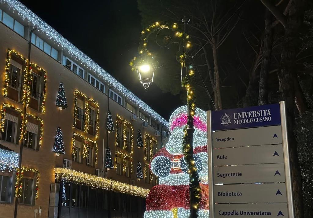 Immagine notturna del campus dell'Università Niccolò Cusano decorata per le feste, con una grande scultura di Babbo Natale in luce e un lampione addobbato.