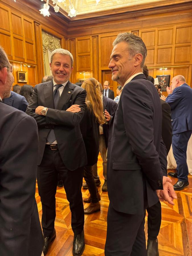 Two men in formal suits smiling and engaging in conversation at a social event in a wooden-paneled room.