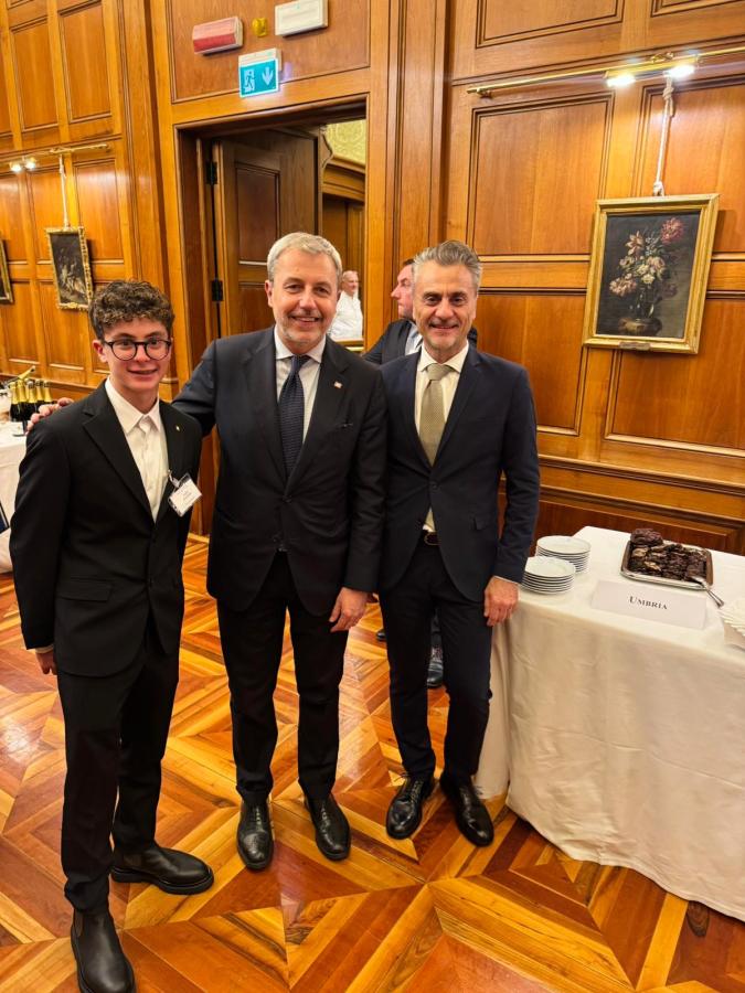 Three men posing together in a formal setting, with wooden paneling in the background. One man on the left wears glasses and a black suit, while the man in the middle is in a dark suit with a tie. The man on the right wears a light-colored suit and tie. A table with desserts is visible in the background.
