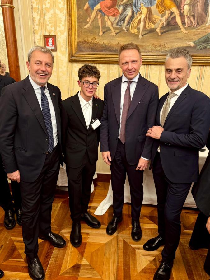 Four men posing together for a photo in a formal setting, with one young man wearing glasses in a black suit and three others in business attire. A decorative painting is visible in the background.