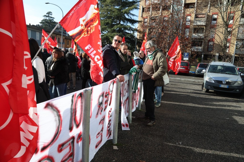 A protest scene with people holding red flags and banners, some individuals engaged in conversation, and a backdrop of buildings and parked cars.