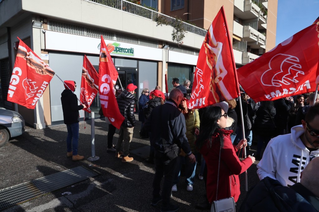 A group of protesters holding red flags with the CGIL logo outside a building. The scene includes people wearing winter clothing and hats, signifying a demonstration.