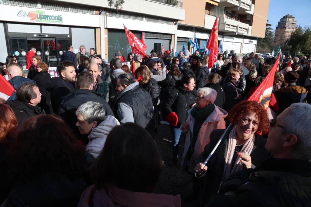 A large group of people gathered in a protest, holding flags and banners outside a storefront. The scene shows a diverse crowd engaging in conversation, with some individuals appearing animated and smiling.