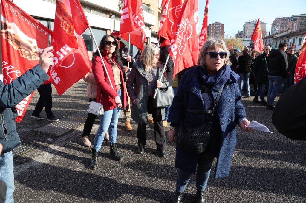 A diverse group of people holding red flags and participating in a protest. One woman in front is speaking, while others listen and hold signs, creating a scene of solidarity and activism.