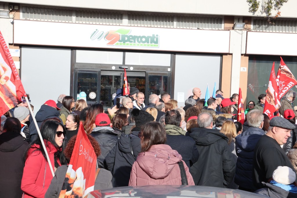 A large crowd of people gathered outside a supermarket, holding flags and banners, demonstrating or protesting. The scene shows a diverse group of individuals with varying expressions and attire.