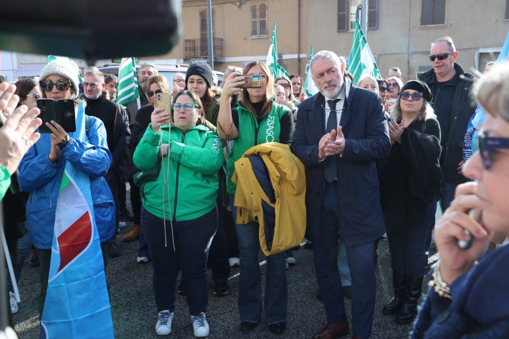 A crowd of people gathered at an outdoor event, some holding flags. Several attendees are taking photos with smartphones, while others are clapping and looking attentively towards the front.