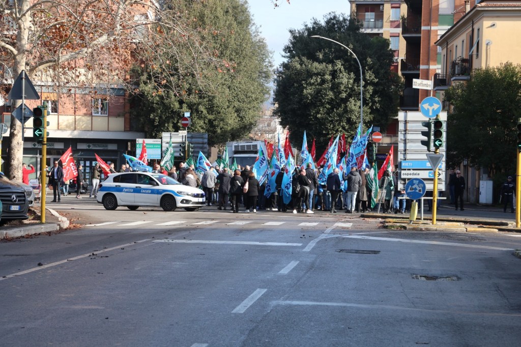 A crowd of people holding colorful flags gathers in the street, while a police car is parked nearby. The setting includes buildings and trees in the background.