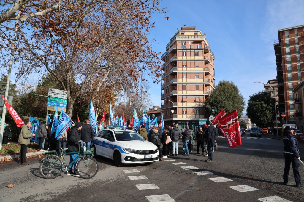 A group of people holding flags gathered on a city street, with a police car parked nearby and residential buildings in the background.