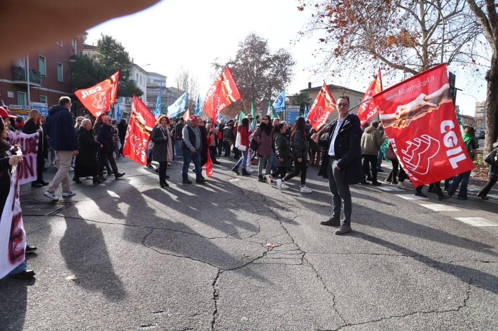 A crowd of people participating in a demonstration, holding red and blue flags. The scene is set on a city street lined with trees, with a mix of individuals dressed in winter clothing.