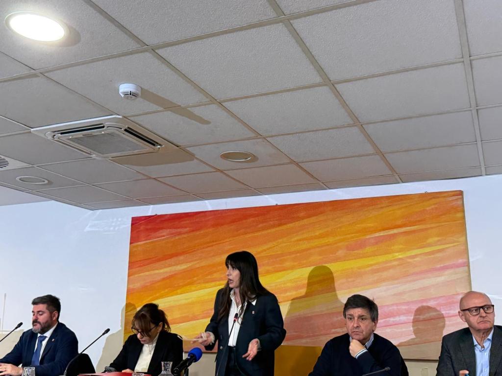A woman standing and speaking at a table with four seated individuals during a conference, with an abstract orange painting in the background.