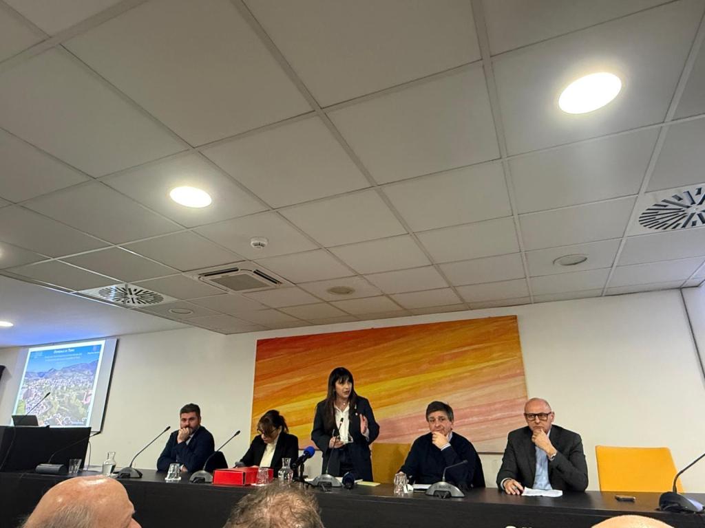 A speaker stands in front of a presentation while three panelists sit at a table in a conference room.
