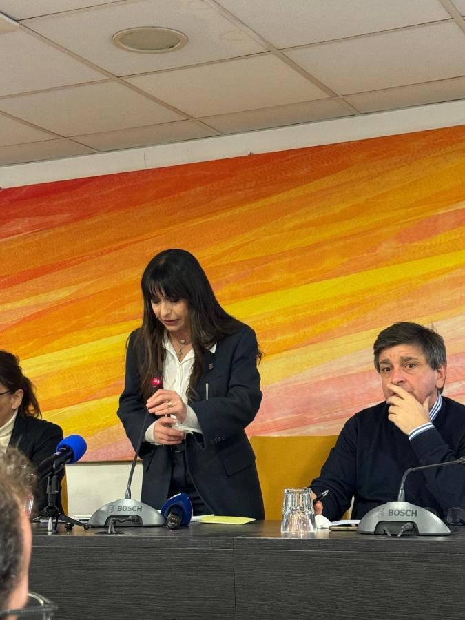 A woman in a black blazer stands at a podium with a red symbol, gesturing with her hands during a press conference. Seated next to her is a man in a dark sweater looking focused, while another woman is partially visible on the left side. The background features an abstract orange and yellow design.