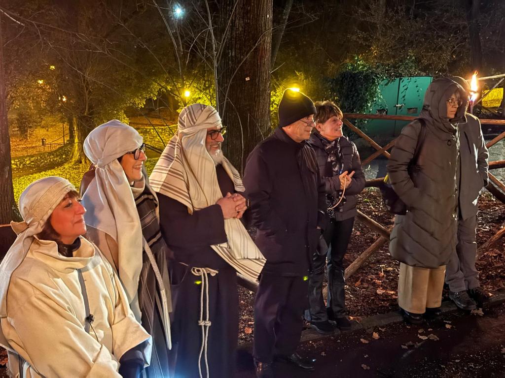 A group of people dressed in traditional attire, gathered outdoors at night, with warm lighting and trees in the background.