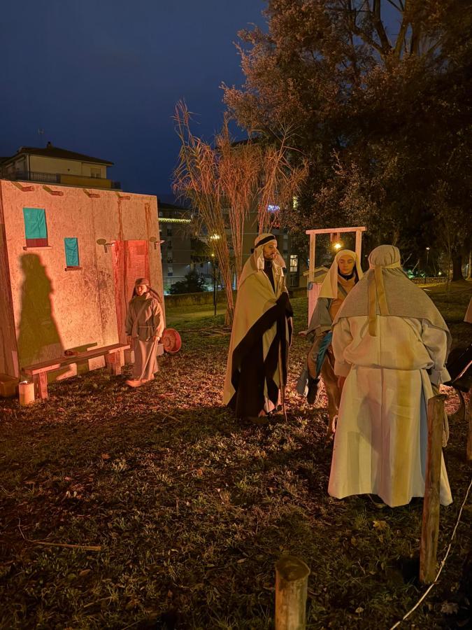 Night scene of actors dressed in historical costumes, gathered near a wooden structure with colorful windows in a park, illuminated by soft lights.