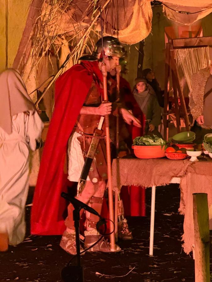 A reenactment scene featuring a Roman soldier in a red cape and helmet, interacting with a table of vegetables in a rustic setting. Other characters in historical attire are present in the background.