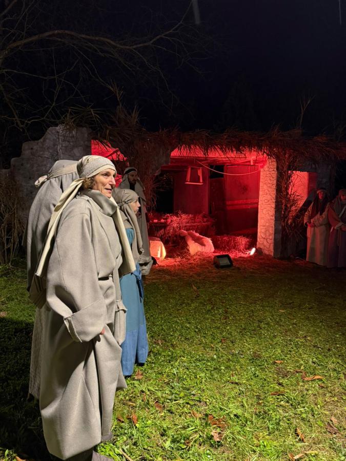 A group of individuals dressed in traditional garments stands in front of a partially illuminated stable, with hay and a warm light creating an authentic atmosphere.
