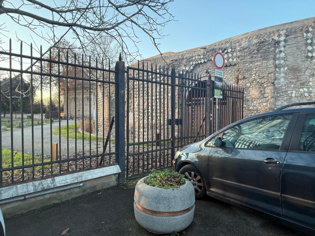 A view through a metal fence showing a stone wall and the corner of a parking area with a parked car and a planter.