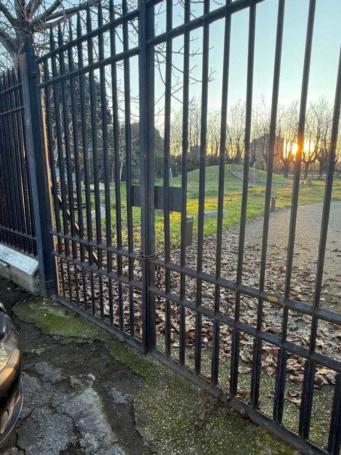 Black metal gate partially open, revealing green grass and a setting sun in the background. The ground is covered with leaves and a parked car is visible in the foreground.