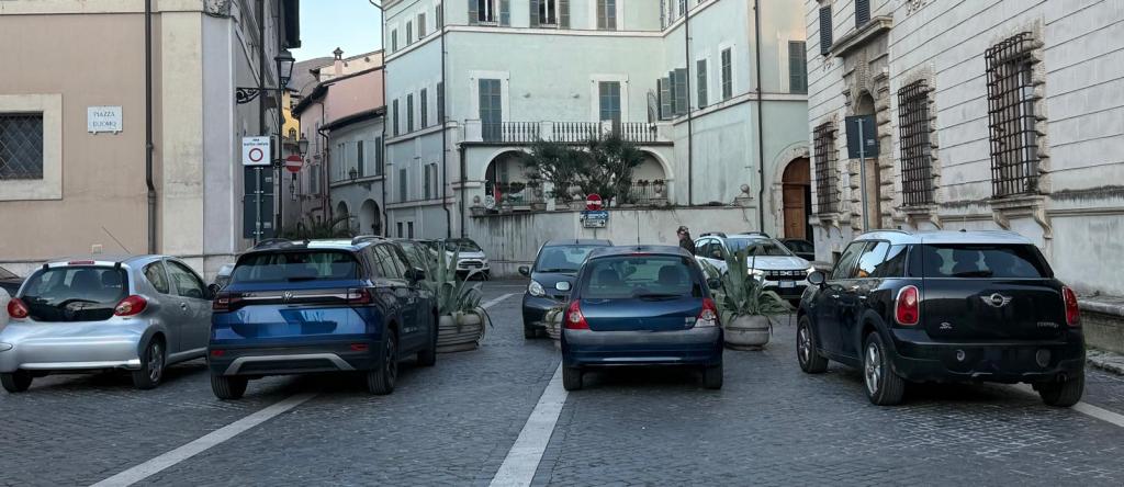 A street scene showing parked cars on a narrow cobblestone street with buildings in the background.
