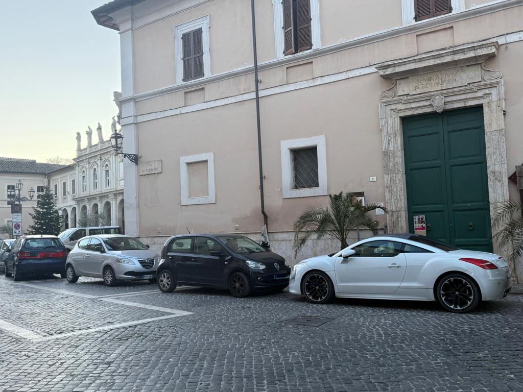 A view of a narrow street in a city with several parked cars, including a white coupe, in front of a building with traditional architecture. The street features cobblestones and a nearby street lamp.