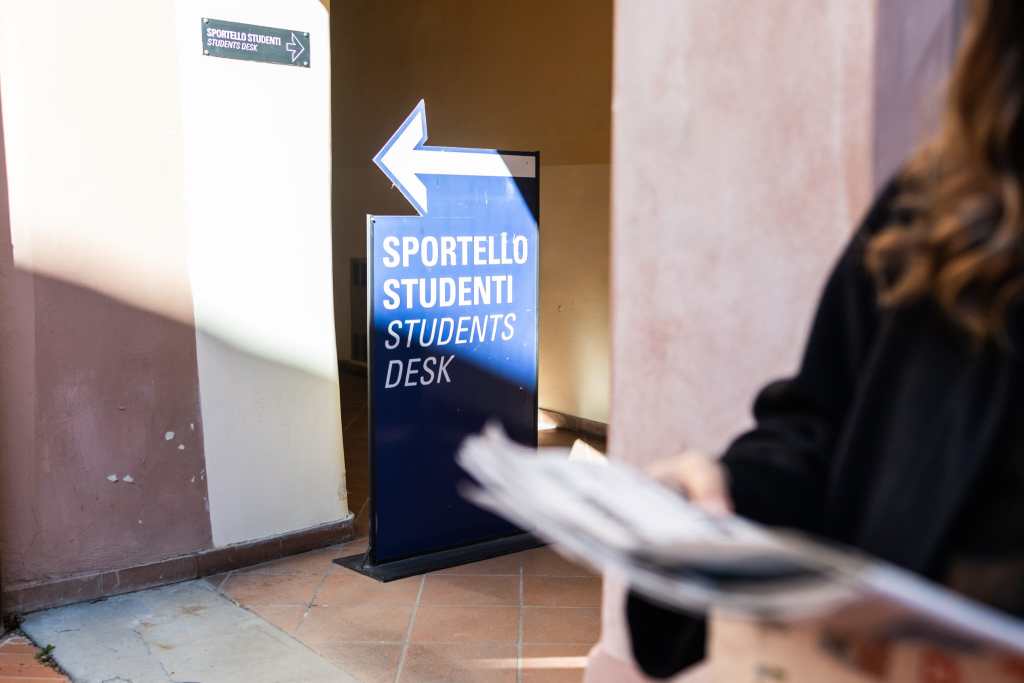 Insegna blu con freccia bianca che indica il 'Sportello Studenti' all'interno di un'istituzione educativa, con una persona che tiene dei documenti in primo piano.