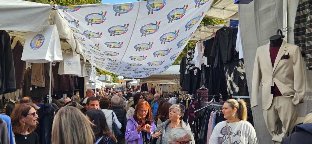 Una vivace scena di un mercato con numerose persone che passeggiano tra le bancarelle, sotto un grande telo decorato con il logo dell'evento. Si possono vedere abiti esposti e un'atmosfera vivace di shopping e socializzazione.