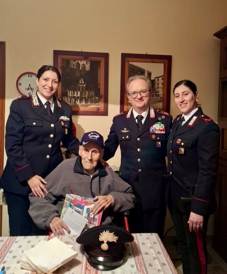 Un vicebrigadiere in congedo circondato da carabinieri in uniforme, mentre riceve un omaggio natalizio, seduto a un tavolo con un berretto e documenti celebrativi.