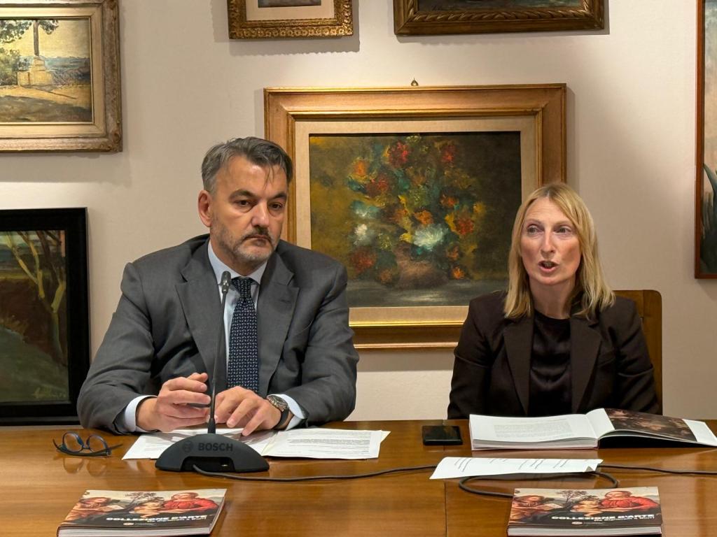 Due relatori seduti a un tavolo durante una presentazione, circondati da opere d'arte appese alle pareti.