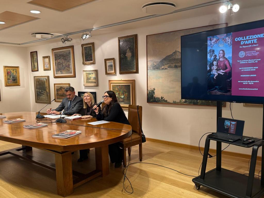 Un gruppo di relatori accanto a un tavolo in una sala d'esposizione, con opere d'arte appese alle pareti e un display che presenta la mostra 'Collezione d'arte. Da Signorelli a Burri'.