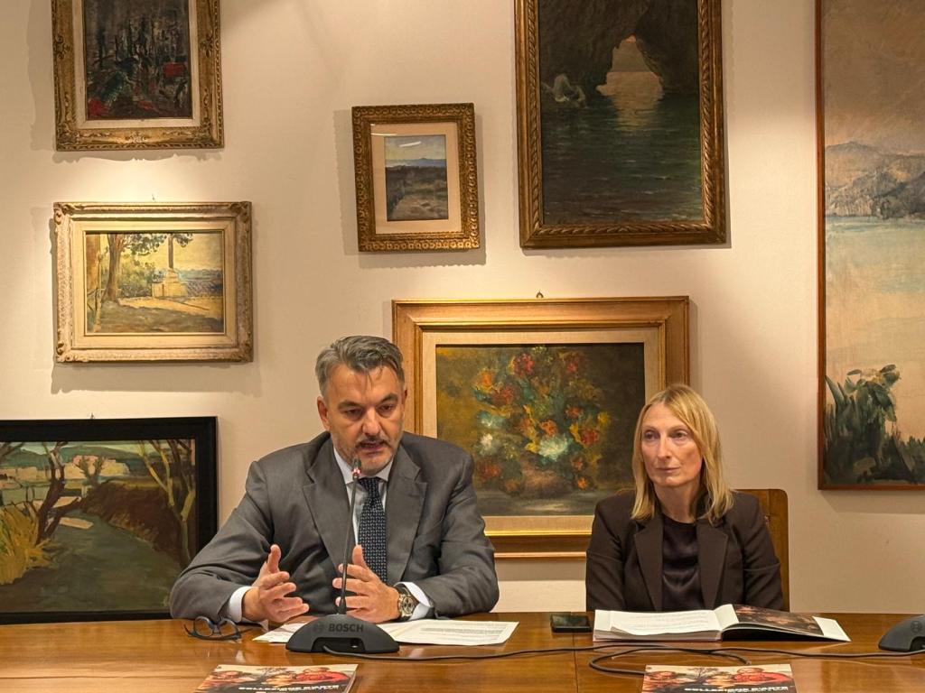 Due persone sedute a un tavolo, circondate da opere d'arte appese al muro, mentre discutono durante un evento culturale.