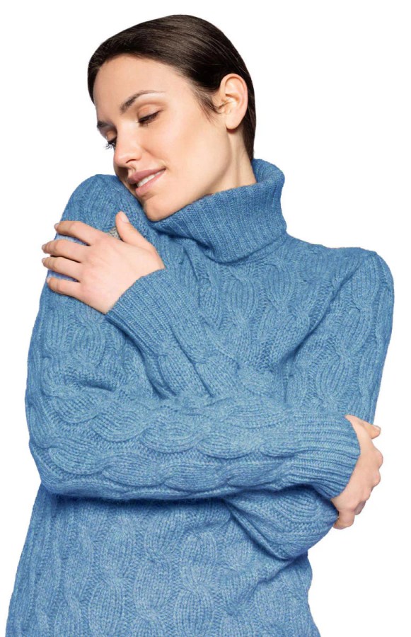 Donna con maglione azzurro a collo alto che abbraccia se stessa, sorridendo e con uno sguardo sereno.