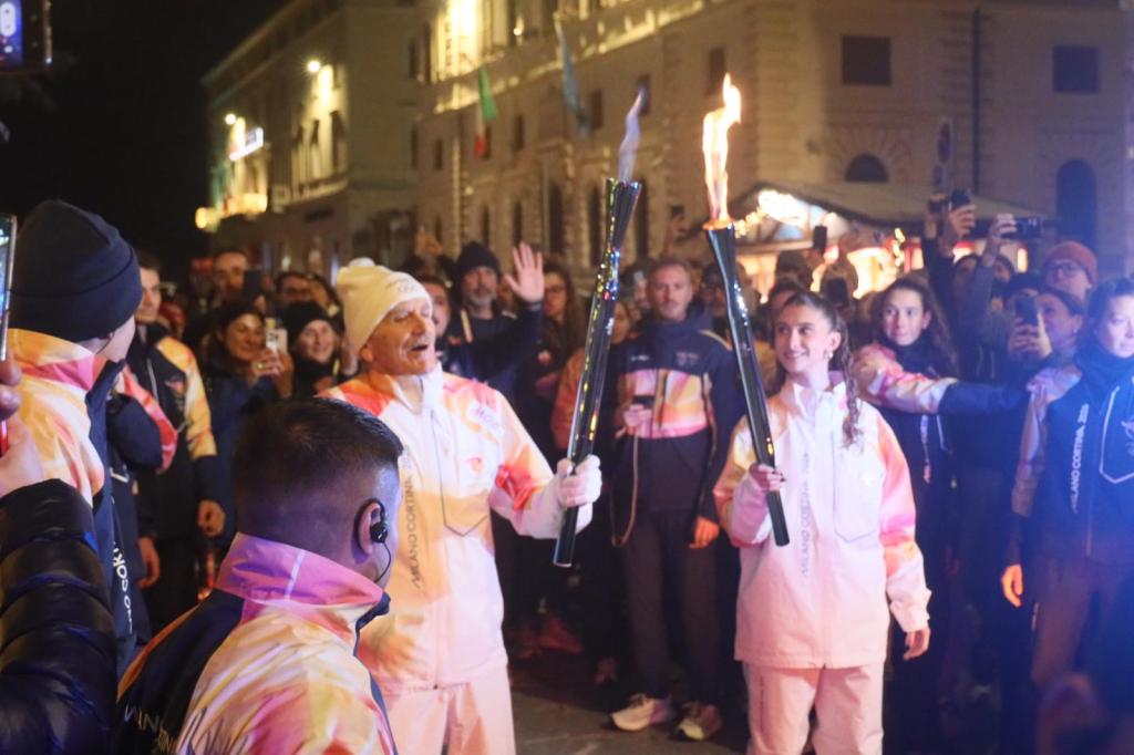 Un momento emozionante della celebrazione dell'arrivo della Fiamma Olimpica a Perugia, con due tedofori che sorreggono la torcia circondati da una folla entusiasta in una piazza illuminata di notte.