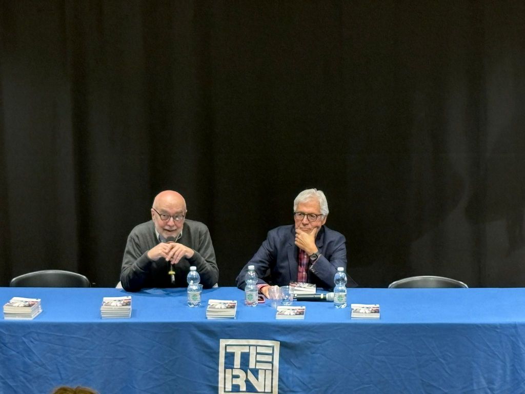 Due uomini seduti a un tavolo durante una presentazione di un libro, con bottiglie d'acqua e copie del libro in vista sul tavolo, di fronte a uno sfondo nero.