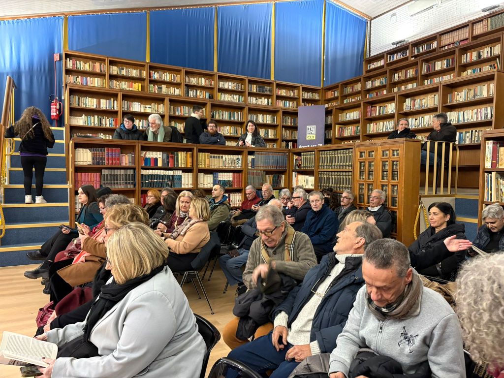 Una sala affollata con persone sedute e in piedi, durante la presentazione del libro "Pensieri nell'acqua" di Franco Fogliano, circondate da scaffali pieni di libri.