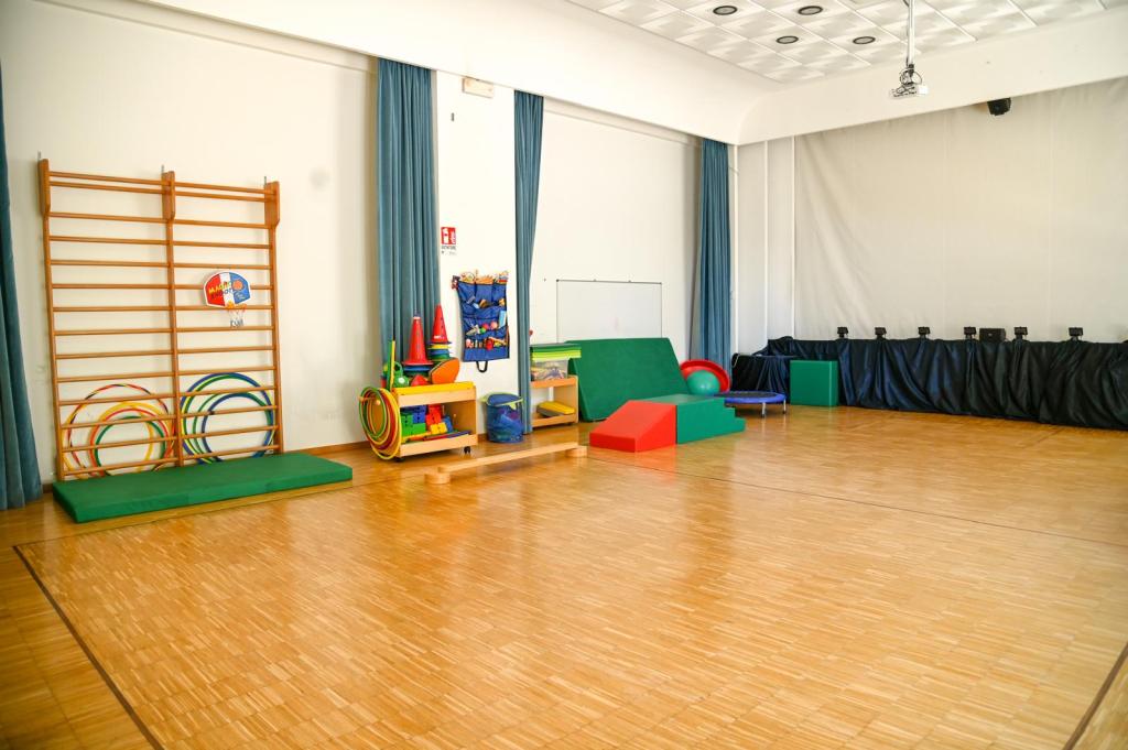 Interno di una sala per attività ludiche per bambini, con attrezzature colorate come un letto elastico, una scaffalatura con giochi e materiali didattici, e tappeti per l'attività fisica.