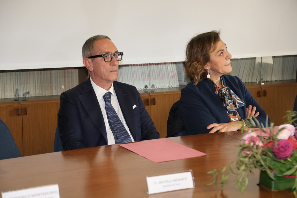 Due relatori durante una conferenza stampa, con un uomo in abito scuro e occhiali e una donna con una sciarpa colorata, seduti a un tavolo con documenti e un mazzo di fiori.