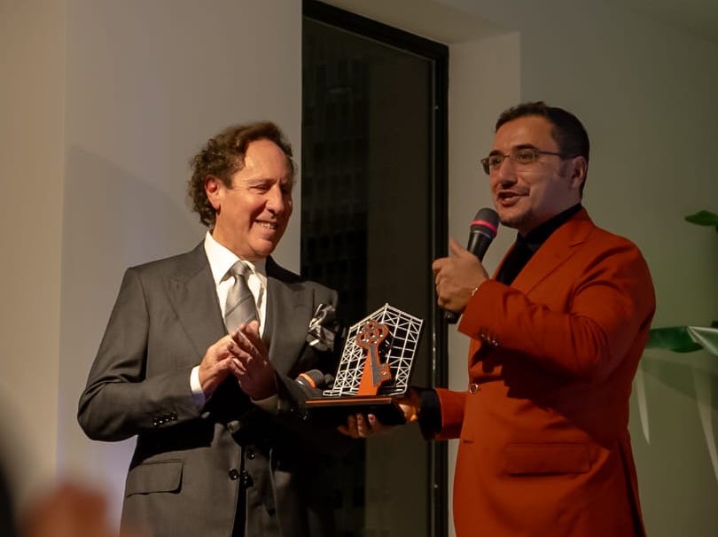 Due uomini si trovano su un palco, uno in abito grigio e l'altro in giacca arancione, mentre il secondo presenta un premio a forma di chiave a quello che indossa abiti più formali.