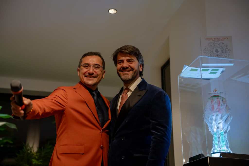 Due uomini sorridenti si trovano in un showroom elegante durante un evento, uno dei quali tiene un microfono. Accanto a loro c'è un trofeo esposto in una teca trasparente.