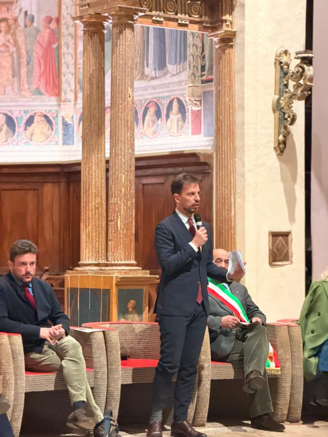 Un uomo in abito formale parla al microfono durante un evento al Complesso museale San Francesco di Montefalco, con dipinti murali e colonne in sfondo; altri partecipanti sono seduti vicino a lui.