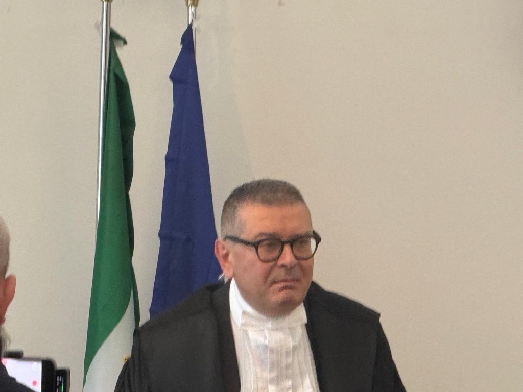 A man wearing a black robe and glasses stands in front of green and blue flags, appearing thoughtful.