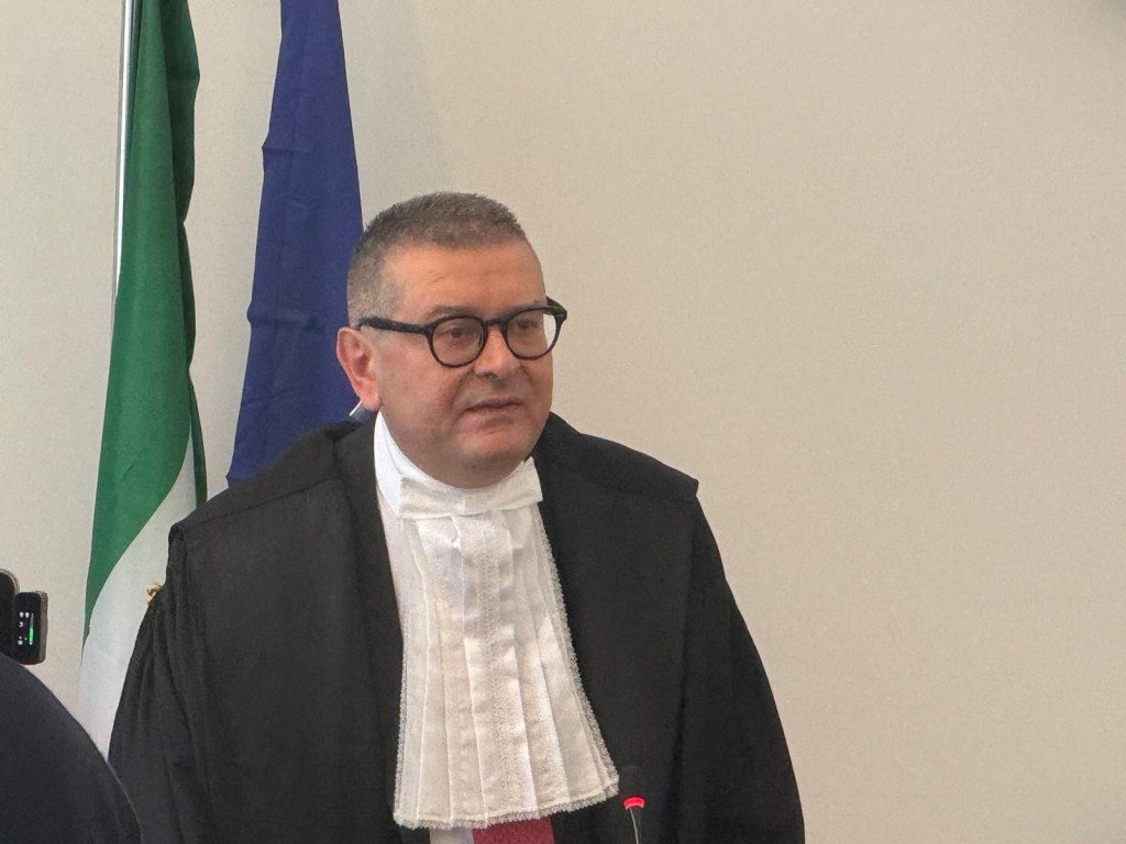 A judge wearing a formal black robe and white collar speaks during a session, with flags in the background.