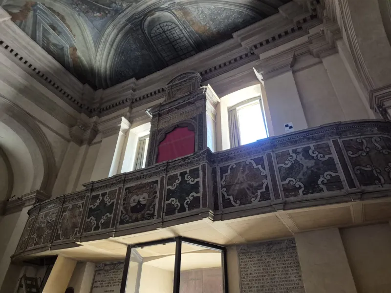 Interior view of a historic building featuring an ornate balcony with decorative elements and a colorful wall mural above. The space is illuminated by natural light coming through large windows.