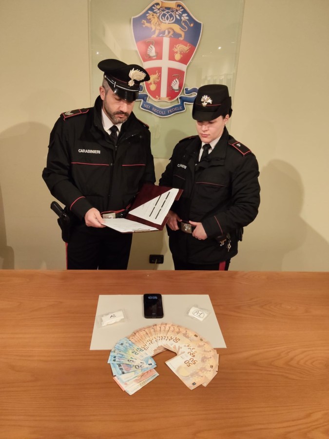 Two Carabinieri officers examining documents while standing at a table with cash arranged in a fan shape, a phone, and two small packages.
